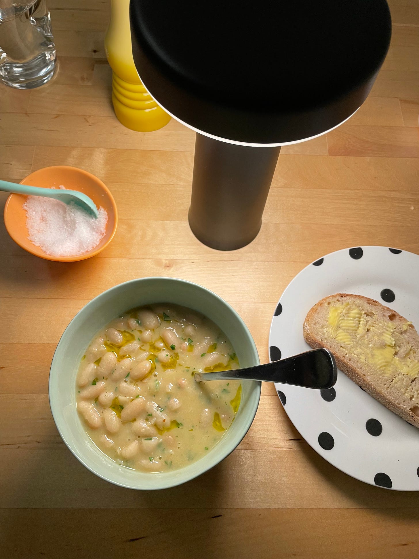 Green bowl filled with cannellini bean soup, a buttered slice of bread and bowl of sea salt nearby.