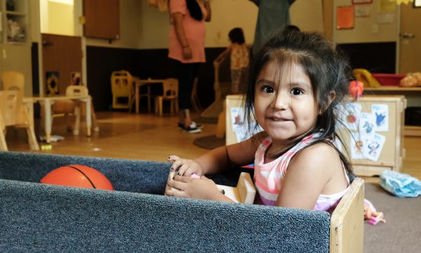 This photo shows a young girl with brown skin and a pink dress sitting in a cart and playing with a basketball in the middle of an early learning classroom.