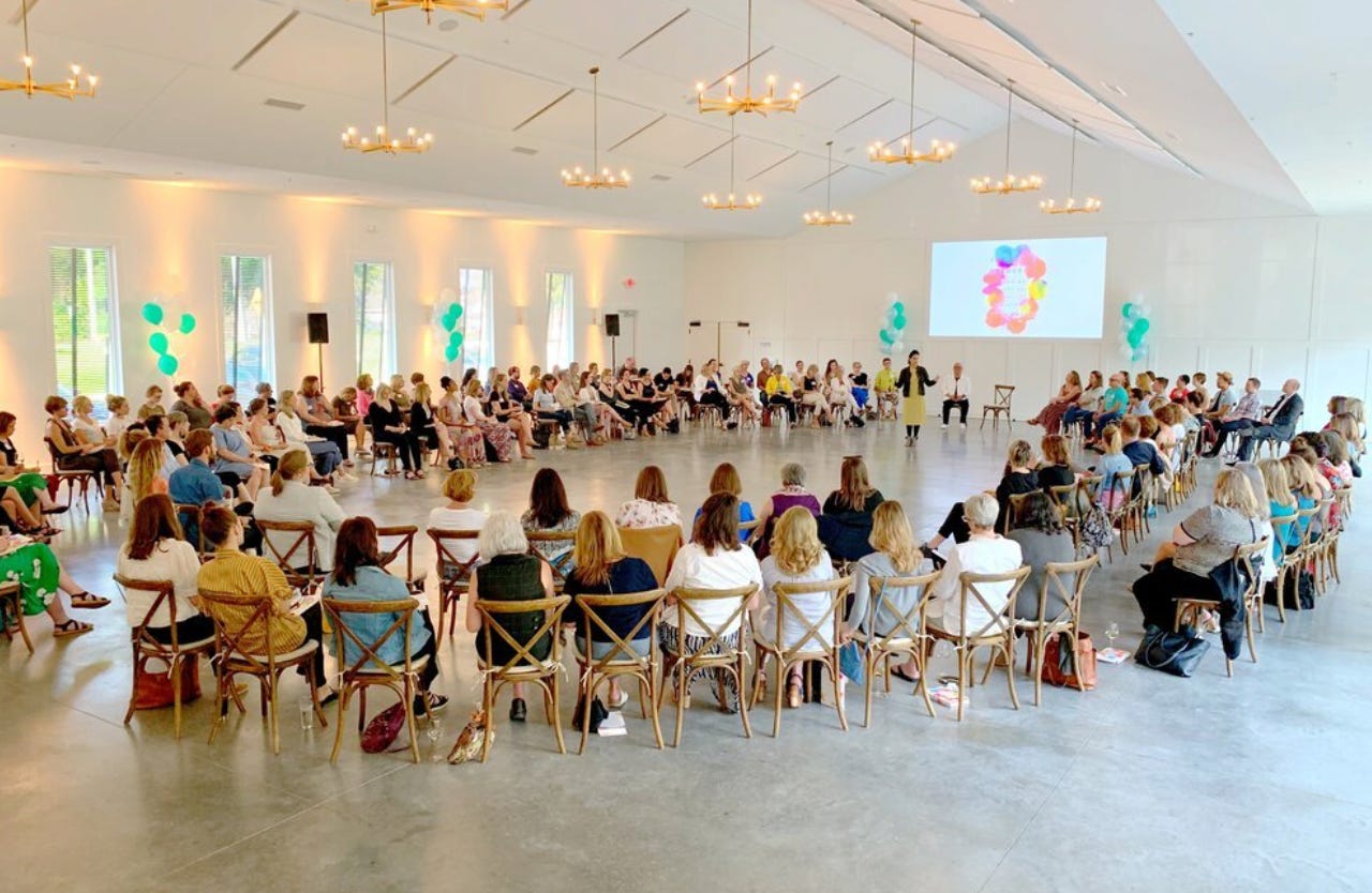 A large group of people all sit in chairs arranged in an oval shape. Priya is standing at the top of the oval, speaking to the group. Behind her, "The Art of Gathering" book cover is projected onto the wall. Everyone's attention is turned toward Priya. 