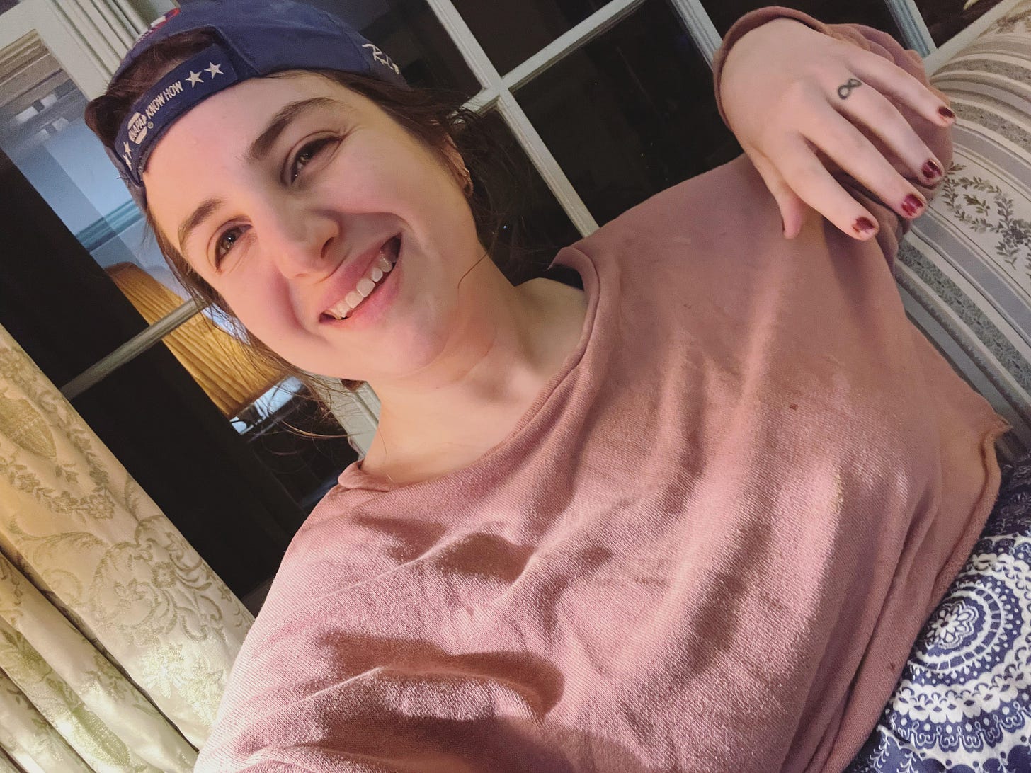 it's jennifer c. martin, a woman in her 30s smiling wearing a ball cap and a cozy pink half sweater and you can see her soft patterned blue pants peakin up. her nails are trash but otherwise she's pretty cute here imo. she's sitting on a couch in front of a window with drapes visible