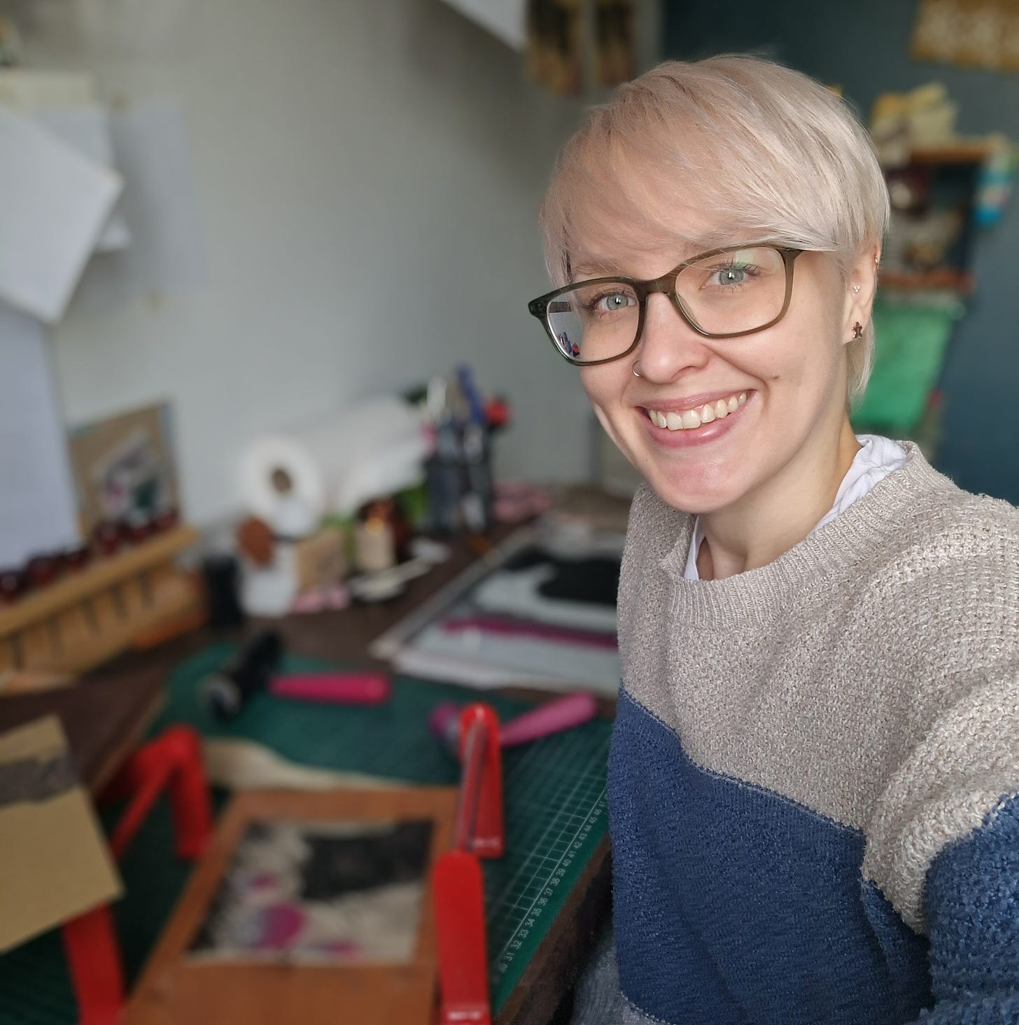 A smiling friendly faced white woman with short cropped blonde hair with a slight peach tone and wearing glasses is staring out at the viewer. She is sitting next to an out of focus work table which has lots of printmakeing equipment on.