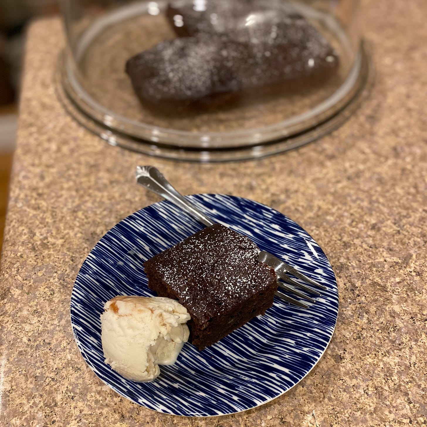 On a small blue and white plate, a square of chocolate cake dusted with powdered sugar next to a scoop of vanilla ice cream with swirls of caramel-coloured syrup in it. A fork rests on the plate, and in the background the rest of the cake sits on a glass cake plate with a cover.