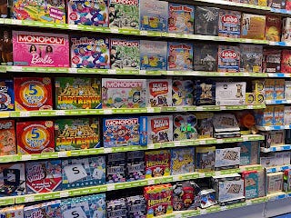 A floor-to-ceiling stocked shelf in an aisle of Smyths toy shop. There are all sorts of games visible including Barbie Monopoly, 5 Second Rule, Scrabble, The Logo Game, Beat the Parents, and Trivial Pursuit.