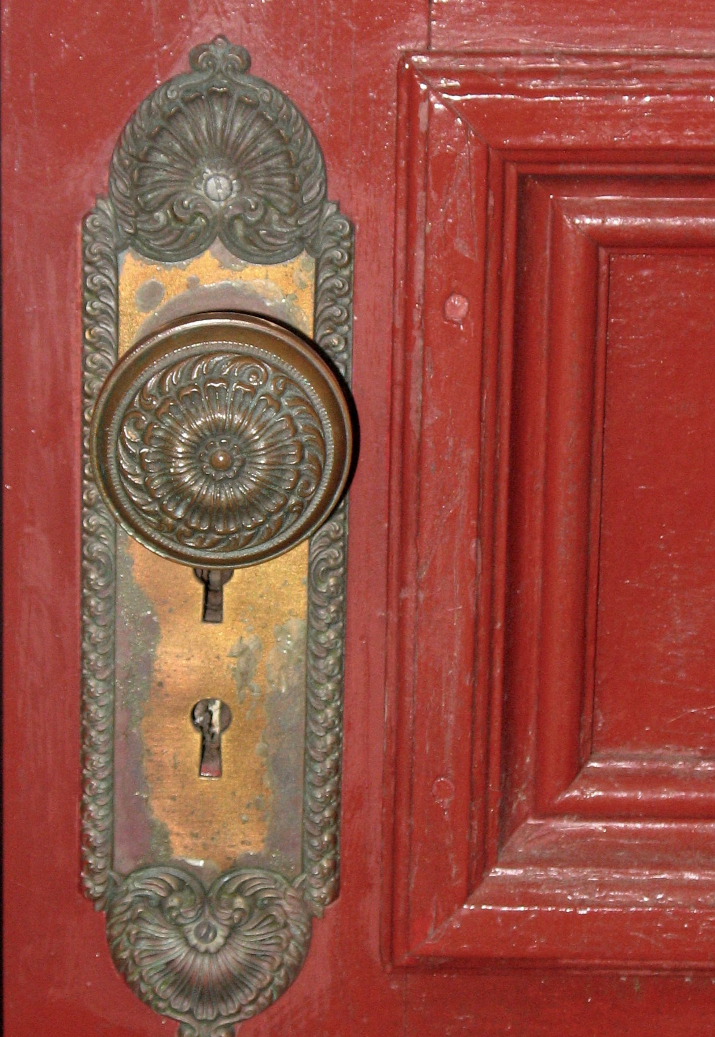 An old wooden door painted red with an intricately designed brass plate and handle. The plate has fan shaped flourishes at the top and a heart shaped one at the bottom.