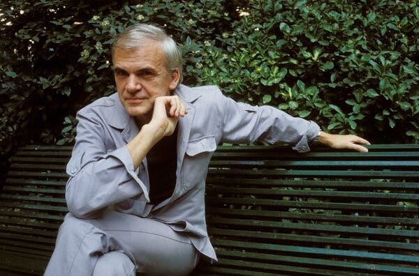 Milan Kundera, in a gray suit, sits on a park bench.