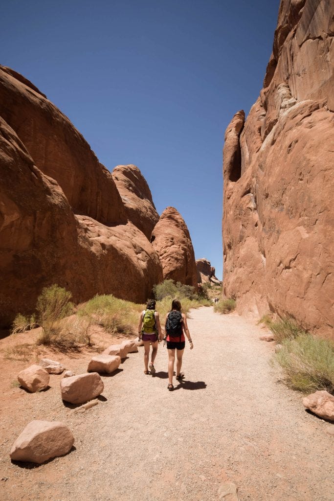 Two Caucasian women hiking through Arches National Park, United States. It is a dry, rocky area with some grass shrubland. The rocks are a reddish colour and tower above the women.