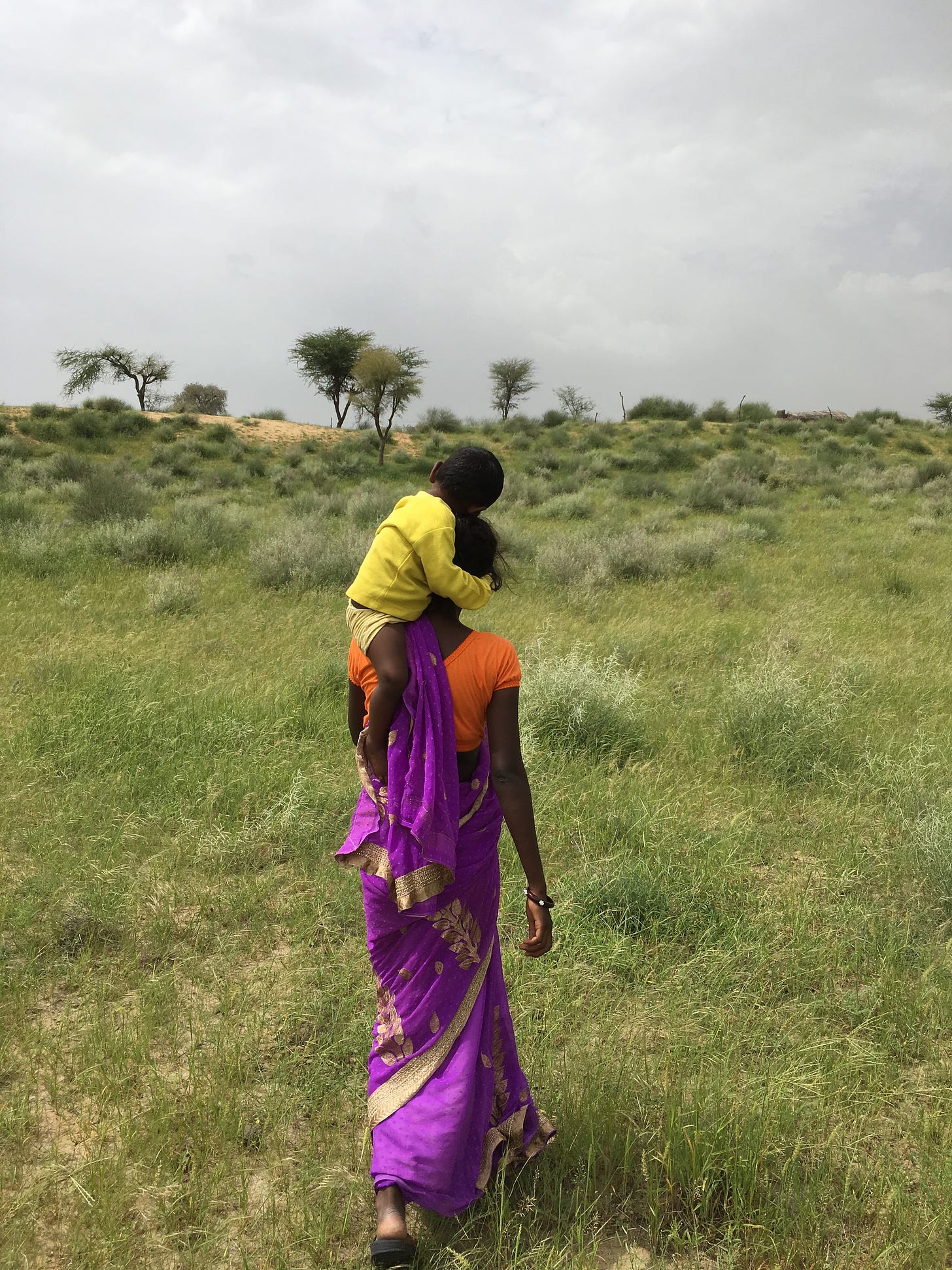 A dark-skinned woman wears a purple and orange saree. She balances a sleeping baby, wearing yellow, on her left neck and shoulder area. They are surrounded by grassy land in a desert. 