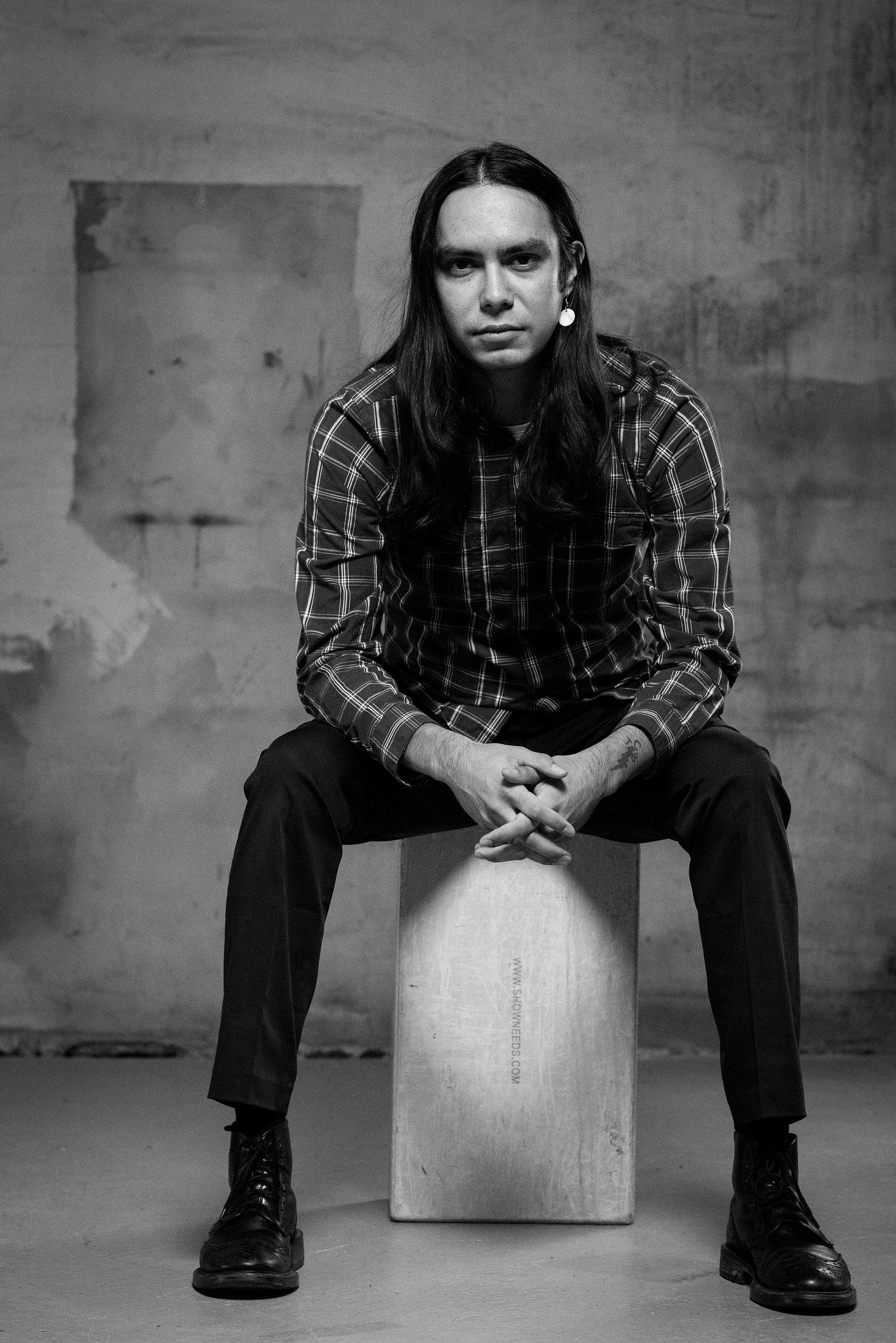 A black and white photo of ishkwaazhe Shane McSauby, a man with long, dark hair, a flannel shirt, and dark pants and boots