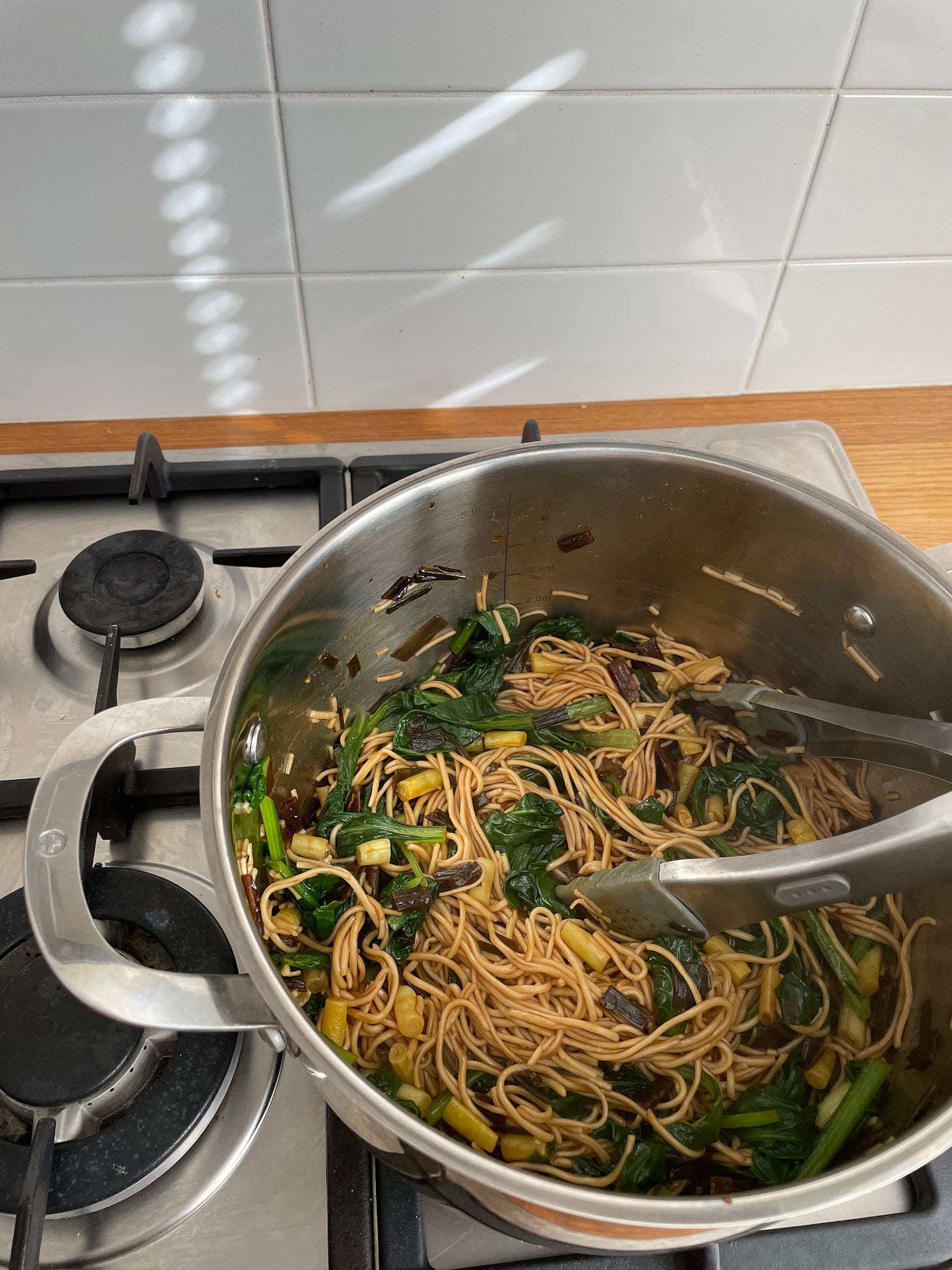 A stainless steel pot filled with egg noodles with chopped beans and scallion oil. It sits on a stove on a sunny evening.