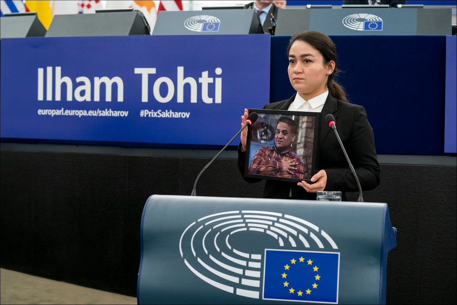 A woman at a podium bearing the EU flag holds an image of a man, and behind her is written the name "Ilham Tohti" in large white letters