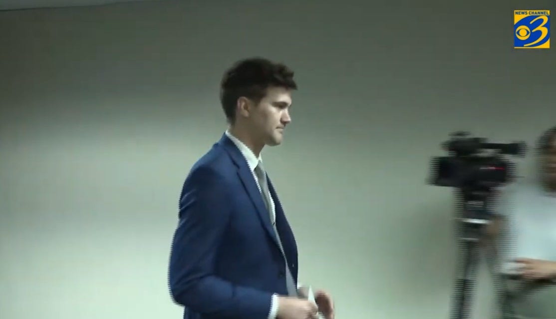Video screenshot of Michigan state Rep. Josh Schriver, wearing a blue suit, walking briskly toward the back of a room, past members of the press. Video screenshot of Michigan state Rep. Josh Schriver, wearing a blue suit, walking briskly toward the back of a room, past members of the press.