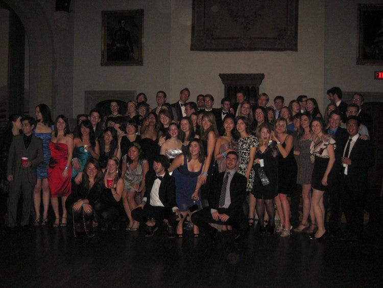 Poorly lit room with group of 50+ students in fancy clothes smiling for the camera.