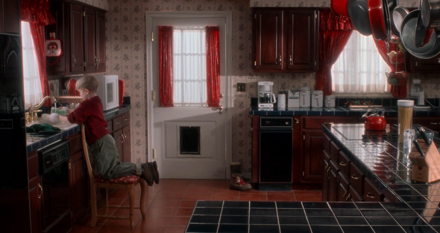 Another angle of the kitchen facing the opposite direction toward the back door. Same deep reddish cabinetry, blue countertops, a gold sink fixture, and red curtains.