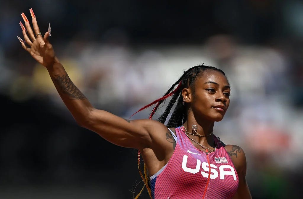 A Black woman with long braids, wearing a pink tank-style track suit, as she's in motion running and raising her right arm up with her hand held up and fingers, with long manicured nails, spread out.