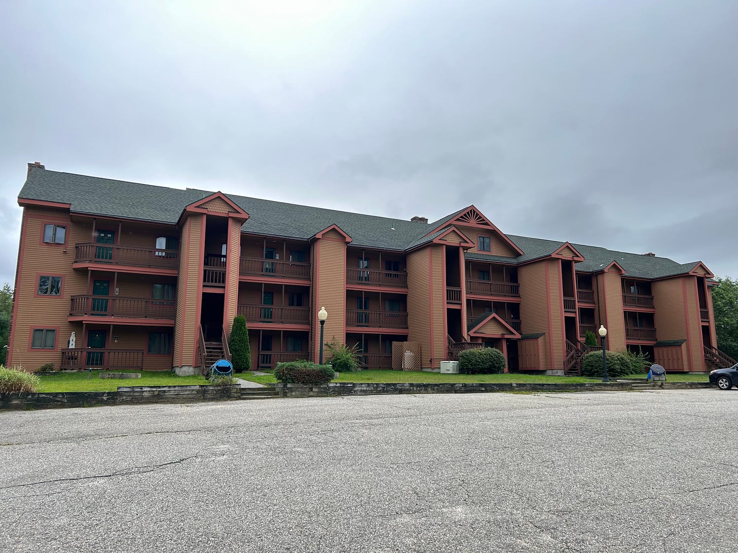 Photo of large three-level condo with brown walls and red trim, no cars in the parking lot, three stairways, overcast skies