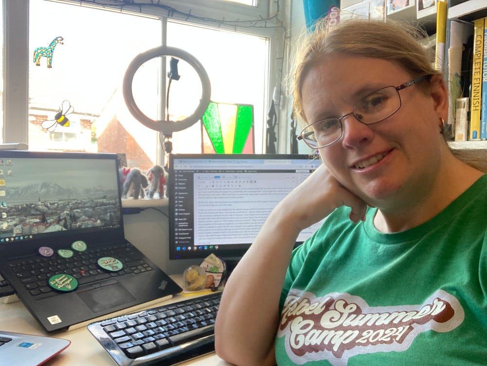 Me, sitting at my desk in my green Rebel Summer Camp t-shirt. Behind me is a laptop with badges scattered over the keyboard and an external monitor, plus a ringlight on the windowsill and my stained glass panel glowing visibly behind the monitor.