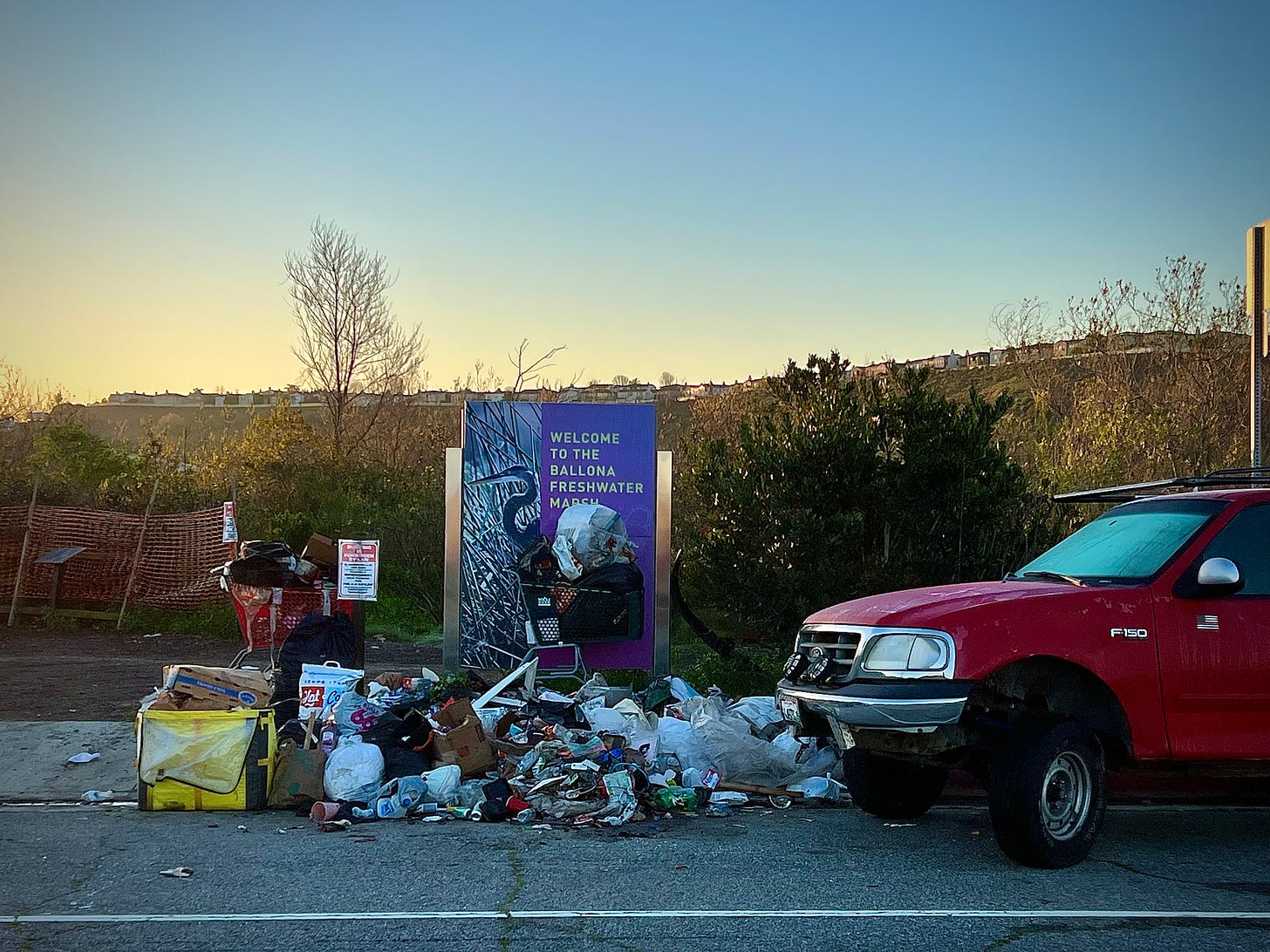 Picture of a sign that says "Welcome to the Ballona Freshwater Marsh," with an image of a great blue heron, in the front of which is a huge pile of trash, two shopping carts loaded with junk, and the front end of a red Ford F150 pickup truck with a frosted windshield and an American flag decal below the drivers' side mirror