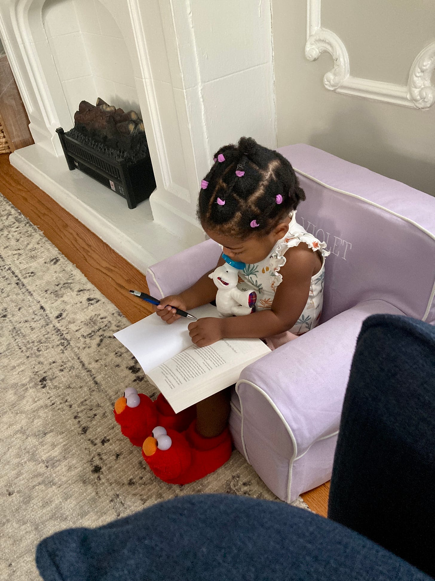 Picture of a Black toddler girl sitting on a kid-sized purple chair with a pen in her hand a book in her lap wearing Elmo slippers