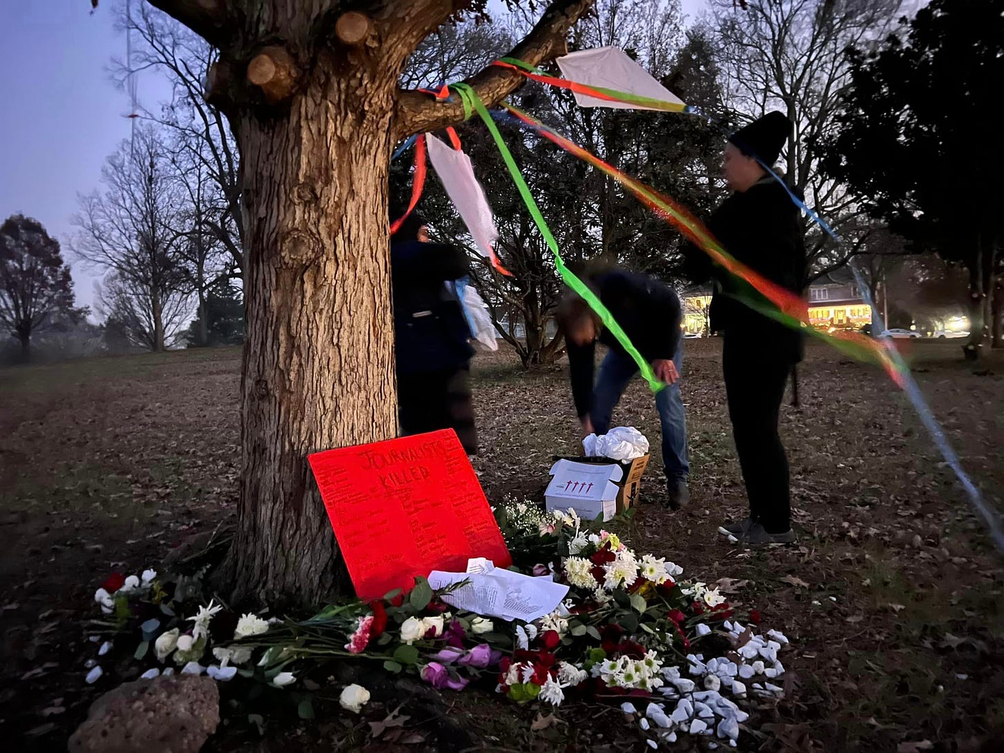 White kites hang from a tree where a sign of journalists killed in Gazas lays, flowers surrounding the tree.