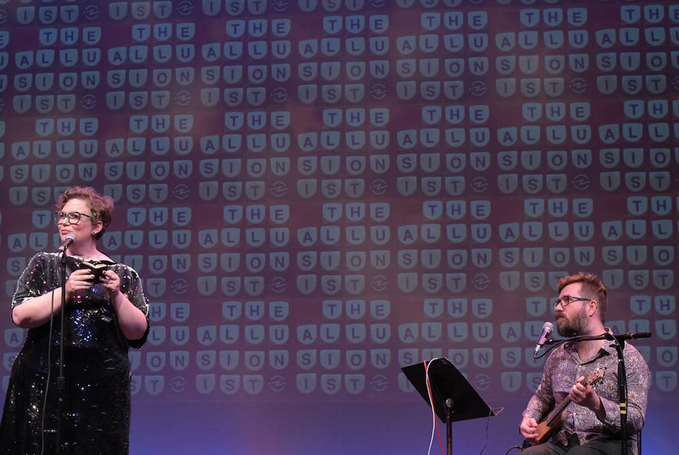 Helen Zaltzman and Martin Austwick are on stage. The backdrop is the Allusionist logo - a repeating pattern of the words 'The allusionist' spelled out in white Boggle dice on the orange plastic Boggle grid. Martin sits behind a music stand, holding a guitar and with a mic pointing towards his face. At the other side of the stage, Helen Zaltzman stands reading from a very tiny dictionary, smaller than the palm of her hand.