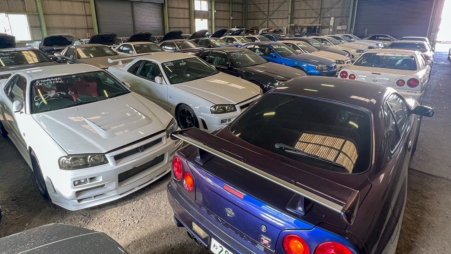 A collection of white, purple and green Nissan R34 Skyline GT-R sports cars crammed into a dimly-lit warehouse in Japan with gray corrugated walls and faded support beams with a gravel floor.