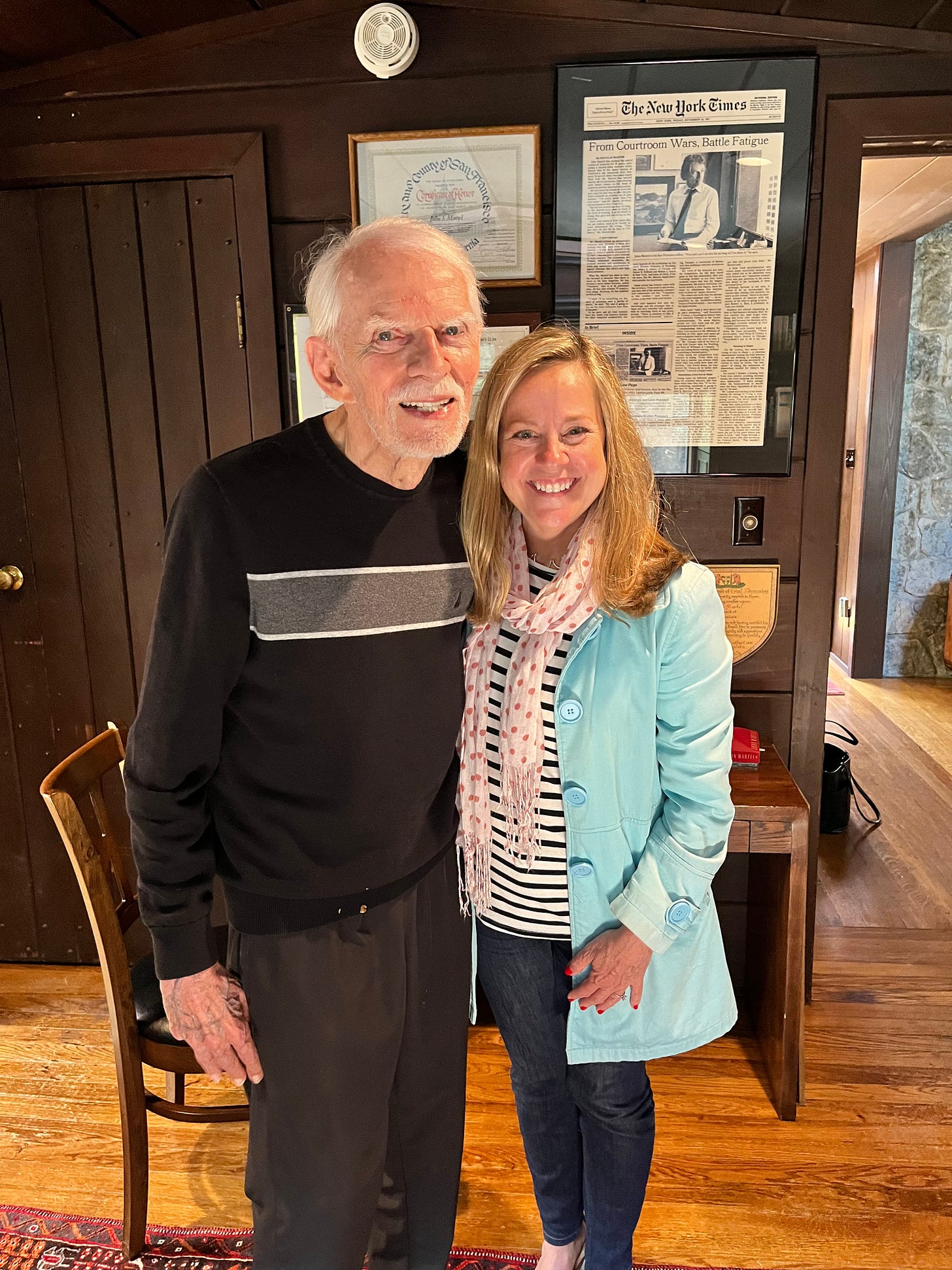 An elderly man and a middle aged woman stand in a wood paneled office in front of framed news clippings and photographs. The man is author John Martel, and the woman is memoir-writing coach and developmental editor Christine Wolf