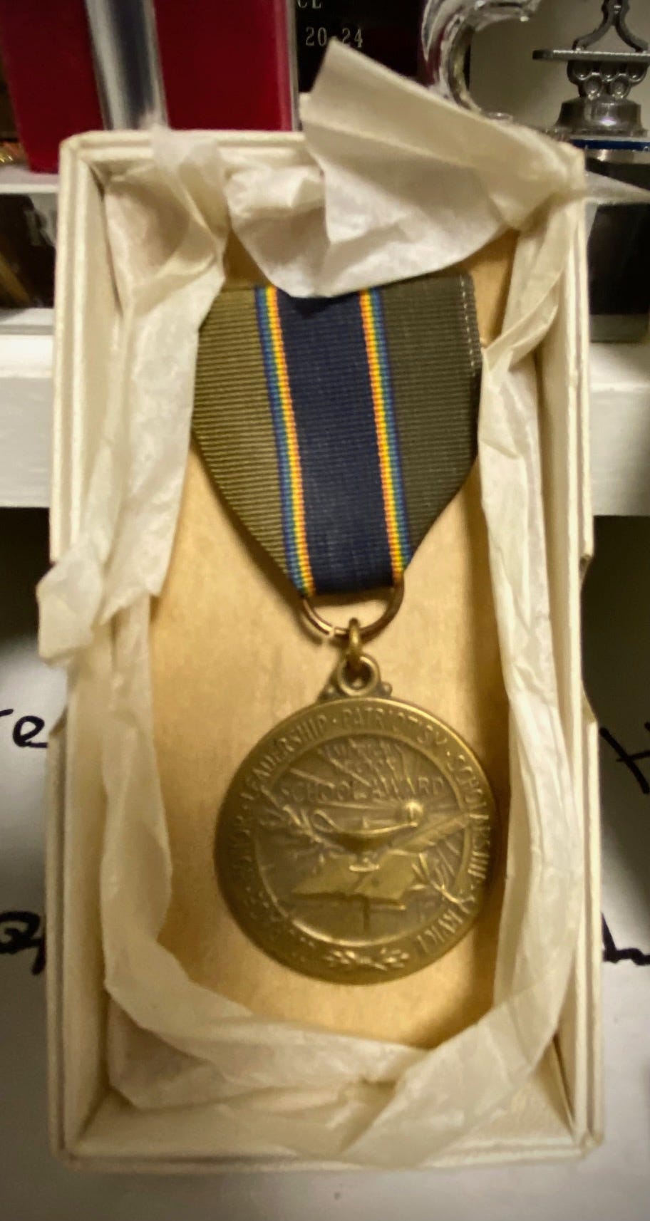 Ribbon with gold medal, nestled in tissue paper in a small box. Inscription on medal reads in center: American Legion School Award. The words in a circle around the edge of the medal read: Patriotism, Scholarship, Service, Courage, Honor, Leadership. Ribbon with gold medal, nestled in tissue paper in a small box. Inscription on medal reads in center: American Legion School Award. The words in a circle around the edge of the medal read: Patriotism, Scholarship, Service, Courage, Honor, Leadership.