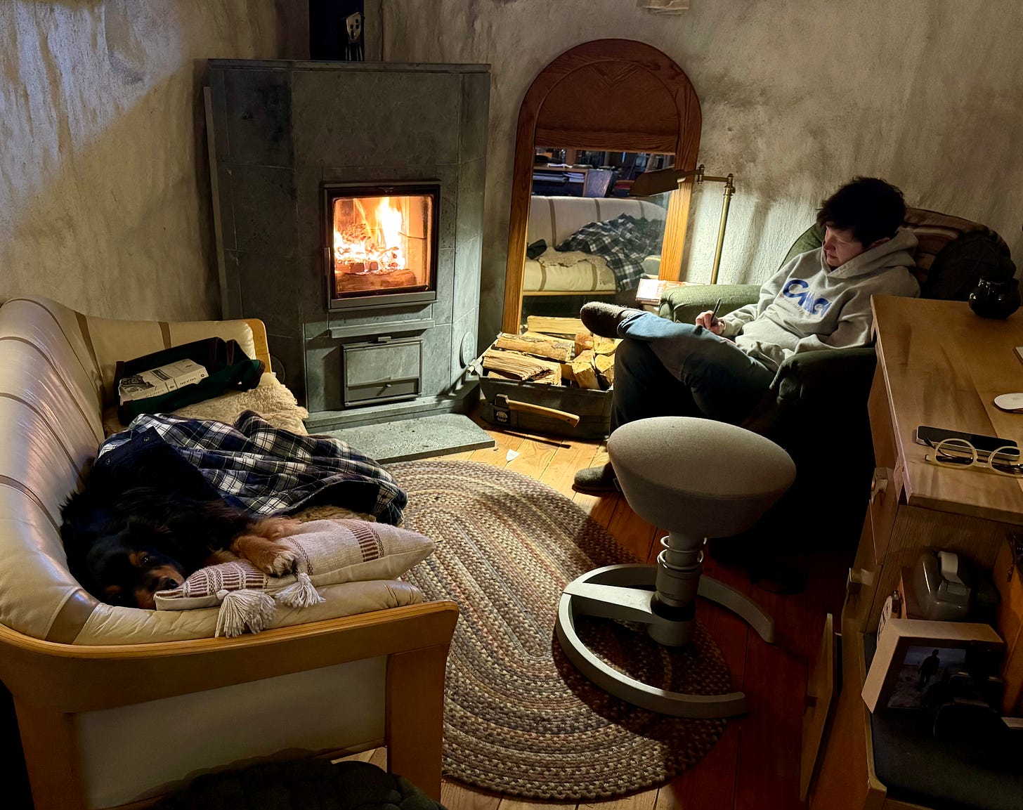 An indoor scene with a person and a dog. The person is seated on a comfy chair, wearing casual clothes and socks, focused on a book, with a pencil in hand. A black and tan dog is lying on the same couch, covered with a plaid flannel shirt, appearing relaxed and comfortable. The room is warmly lit by a wood stove fireplace, with visible flames behind a glass door. There's a round rug on the floor, a stool with a curved base, a wooden desk with various items, including glasses and a smartphone. A large mirror on the right reflects part of the room, enhancing the cozy ambiance.