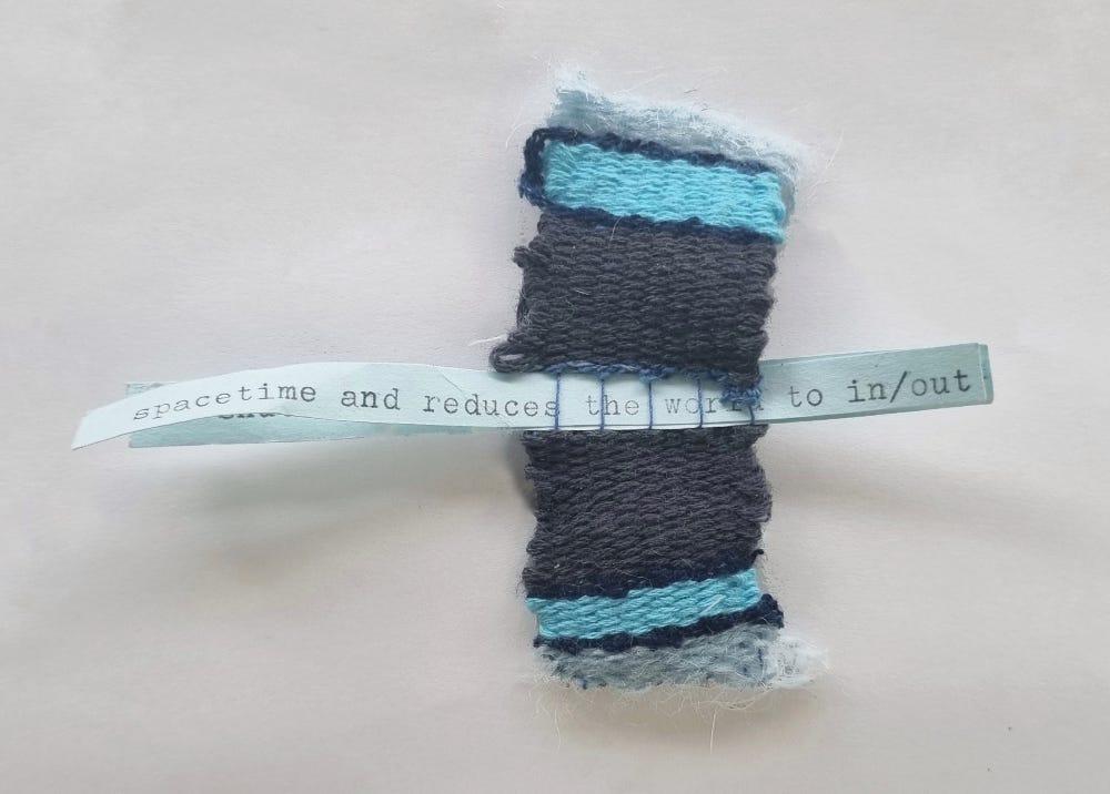 A small, handwoven textile piece in shades of blue and gray. The weaving is rectangular and features different textures and patterns. A strip of paper with typed text is woven through the center, reading "spacetime and reduces the world to in/out".