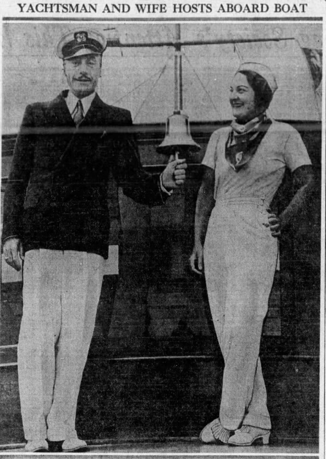 A photograph showing a middle-aged couple. The man stands on the left and is wearing white trousers, a blazer and a nautical captain's hat. The woman, his wife, is wearing a white trouser suit and is smiling broadly as she looks at her husband. Between them there is a bell - the man is holding its rope.