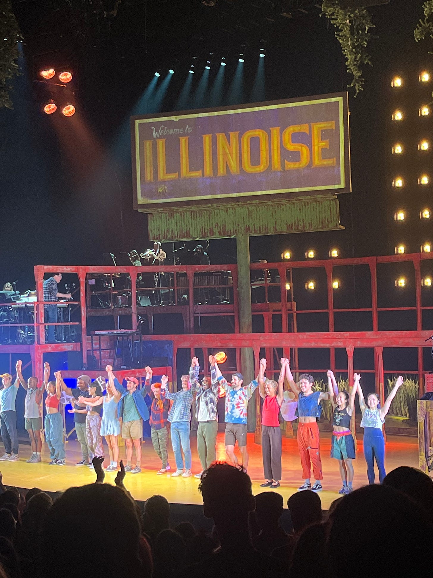 A stage filled with members of the "Illinoise" cast, beneath a billboard sign that says "Welcome to Illinoise," with red structures around the stage