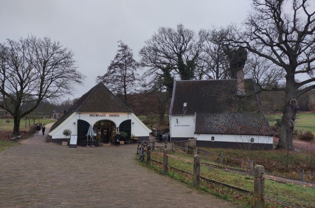 Two buildings with pointy roofs, one holds the cafe and both its barn-style doors are wide open for custom. There are a few trees behind, and a bike is leaning against a low wooden fence in front. The pavement is cobbled.