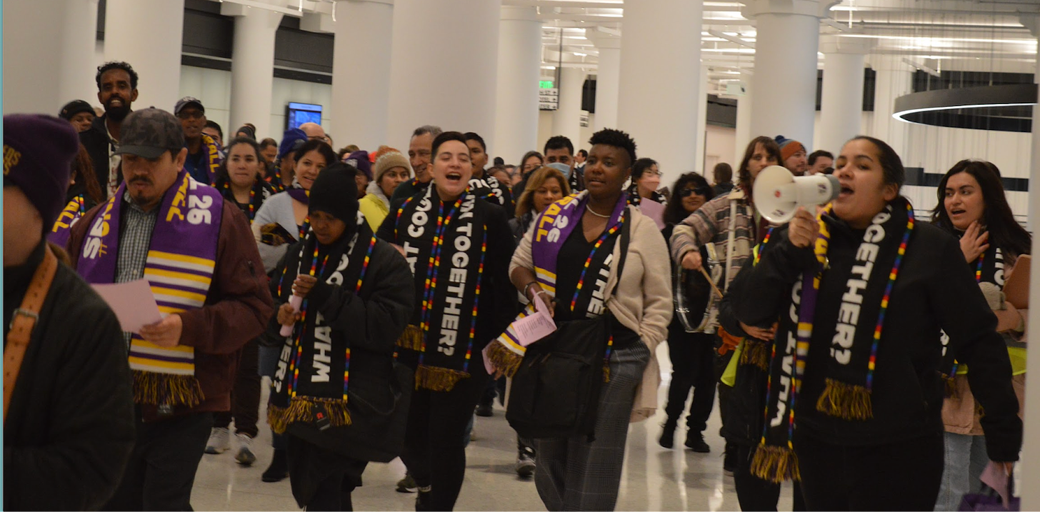 a crowd of people march through a large hallway wearing purple and black scarves reading "what could we win together?"