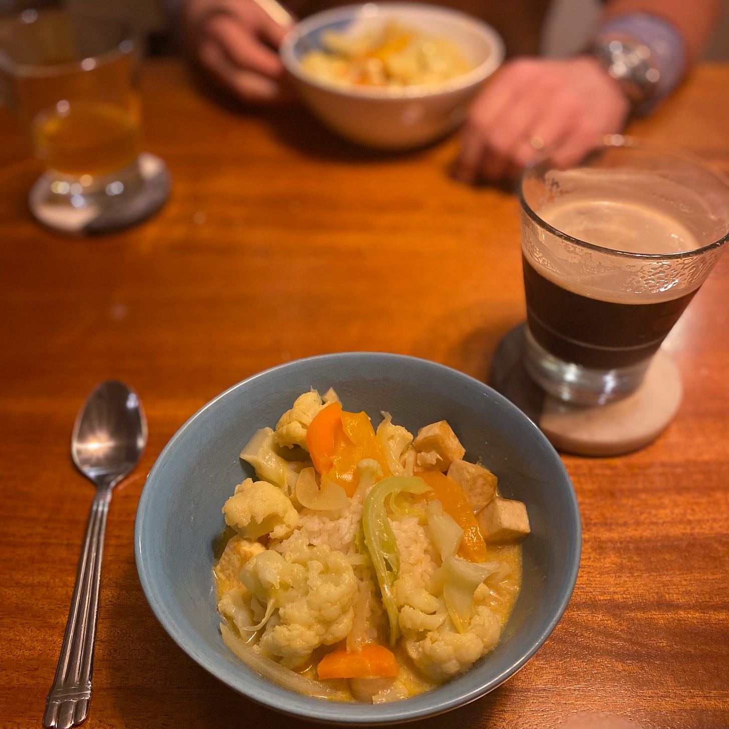 A blue bowl of yellow curry with cauliflower, carrots, and cubes of tofu. Next to it on a coaster is a glass of dark beer. Jeff is across the table with a white bowl of the same, and a glass of lighter beer.