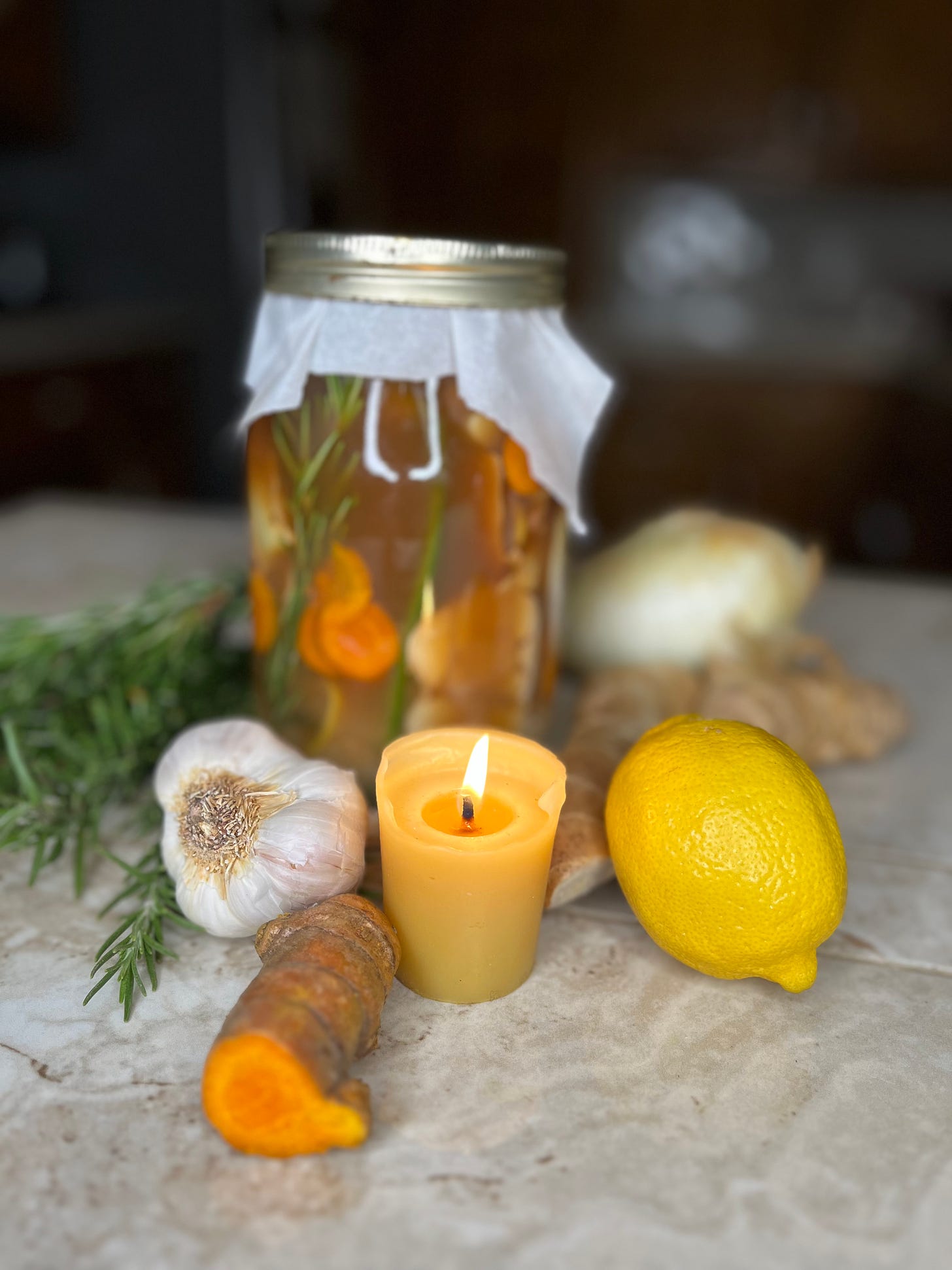 A jar of fire cider with fresh ingredients- rosemary, garlic, turmeric, lemon, onion, and ginger- scattered around it. A lit candle in front of the jar.