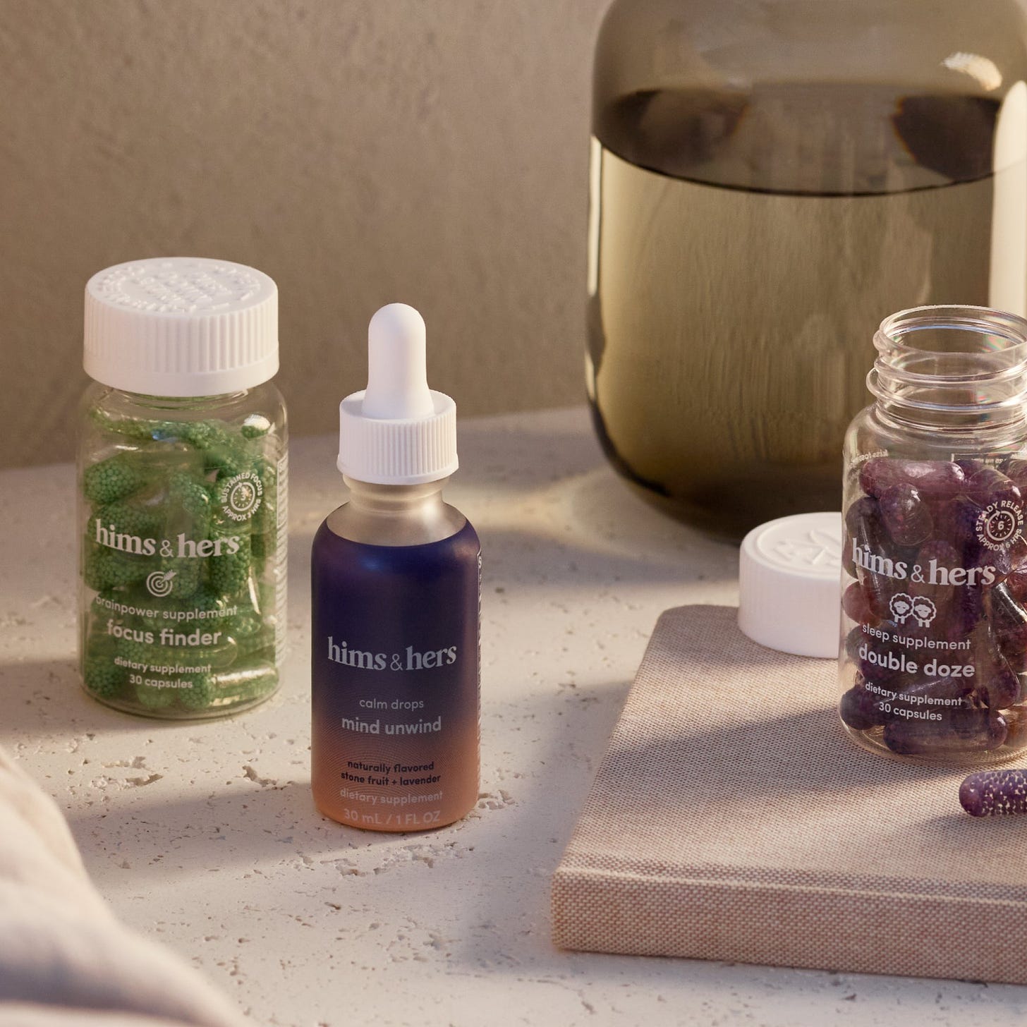 Close-up of Hims & Hers wellness supplements, including Focus Finder brainpower capsules, Mind Unwind calm drops, and Double Doze sleep capsules on a table, showcasing premium health and wellness products for mental focus, relaxation, and sleep support.