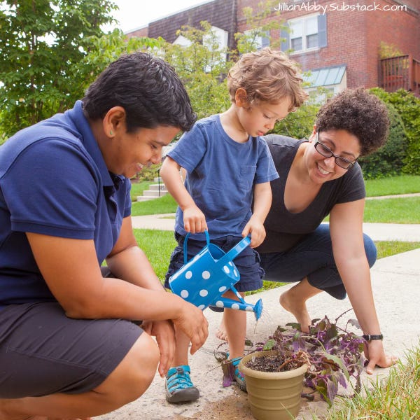 Queer couple crouch to each side of their toddler as he waters a potted plant with a blue watering can.