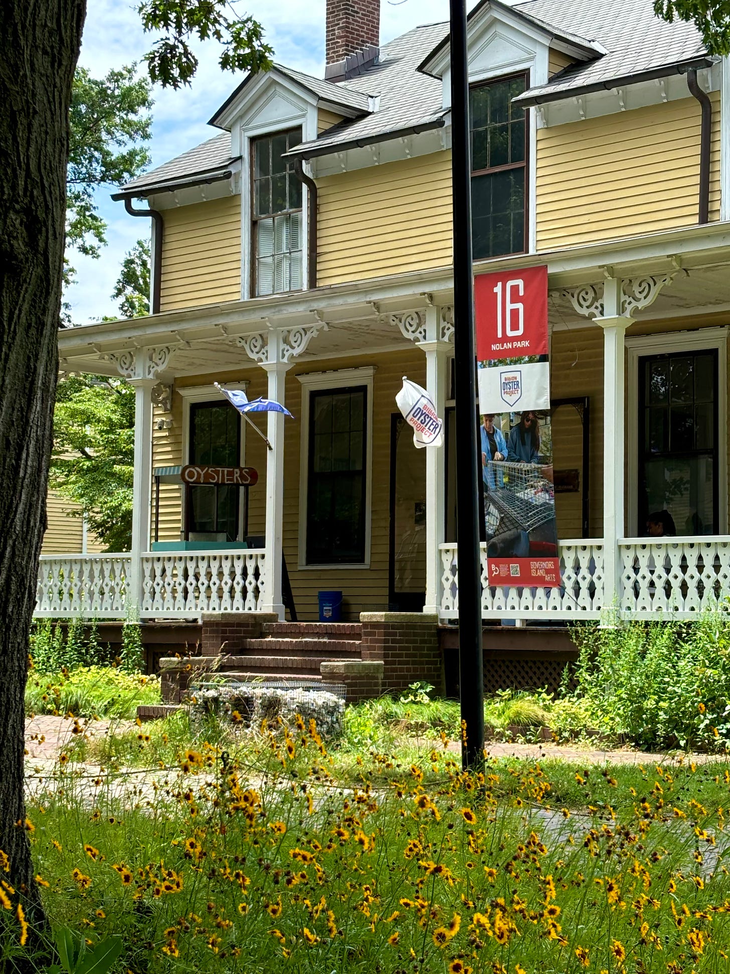 A yellow house with an oyster cart on the porch. There is a tree to the side and yellow wild flowers in front. The entrance is framed by a Billion Oyster Project flag and its address on Governors Island.