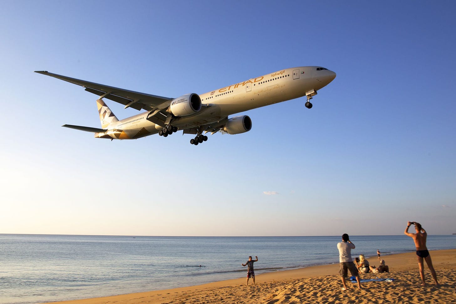 An Etihad Airways airplane flying low over a beach with people watching and taking photos.