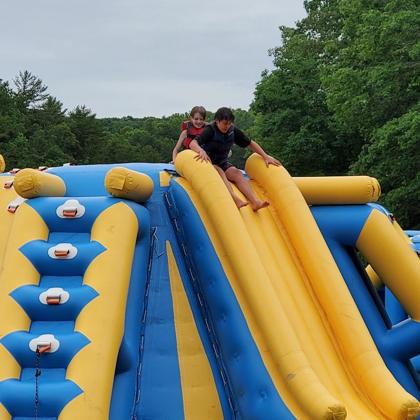 Two white boys in bathing suits and life jackets slide down an inflatable lake toy. 
