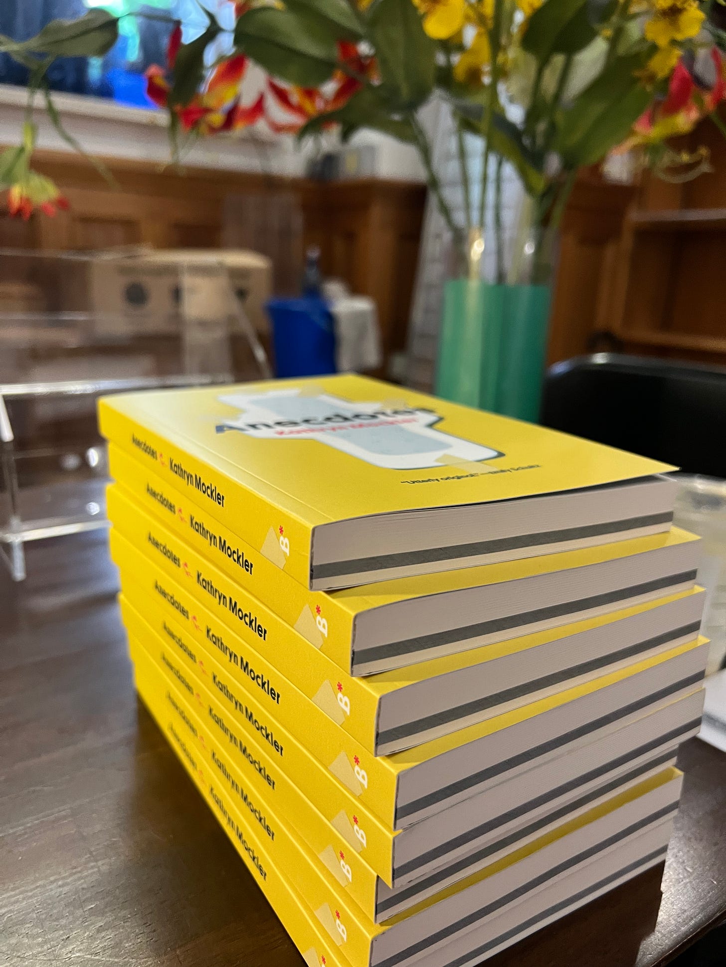 A stack of yellow books with a maxi pad on the cover: Anecdotes by Kathryn Mockler. The books sit beside a bouquet of flowers.