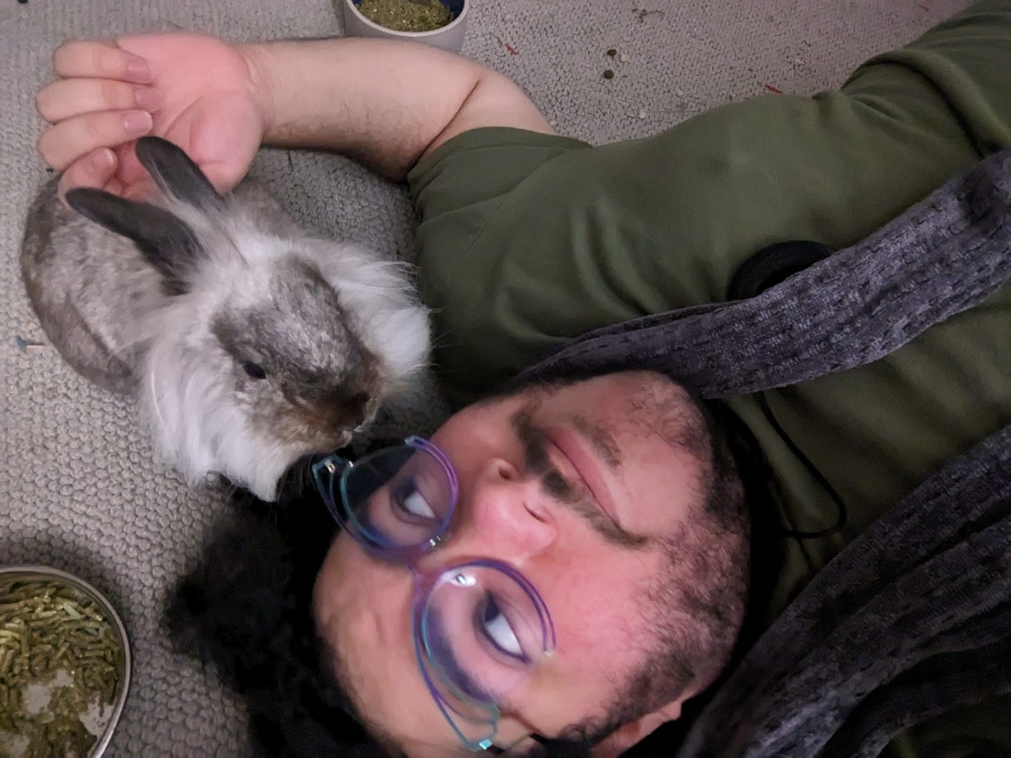 A person (me) lying down on the floor with a grey/white lionhead rabbit whose face is close to mine, and I have the back of my hand on his back. I am looking over at him. Bunny food bowls are in the photo