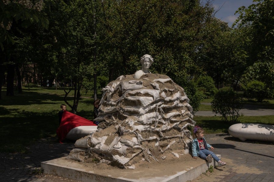 Children play next to a monument covered with sand bags that are falling apart.