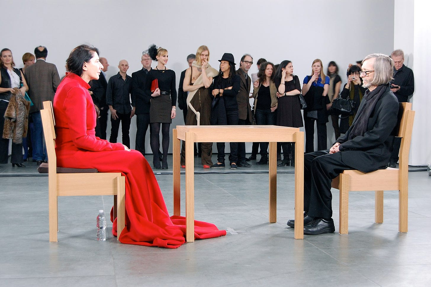 A woman (Marina Abramović) sits in a wooden chair at a small wooden table, gazing at a man who sits in a matching chair in the same position, gazing back.