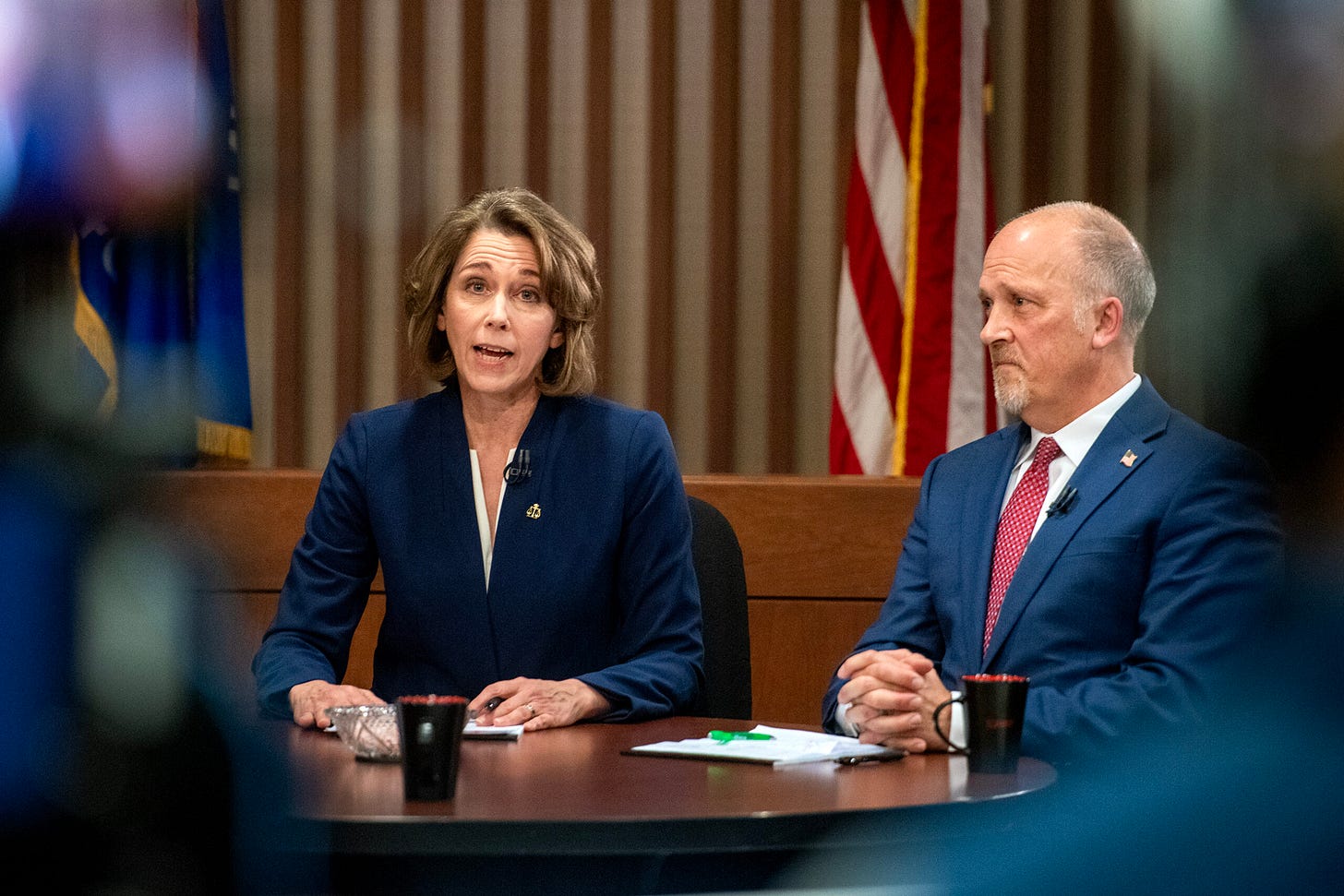 A woman and a man in formal attire sit at a table with microphones and papers in front of a backdrop featuring flags. A woman and a man in formal attire sit at a table with microphones and papers in front of a backdrop featuring flags.