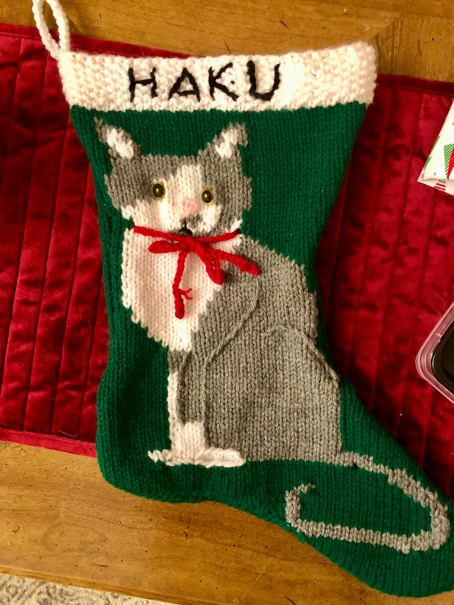 A knitted, green stocking with a white strip at the top and the name "Haku" written in black yarn. The stocking has a picture of a grey and white tuxedo cat wearing a red bow around its neck.