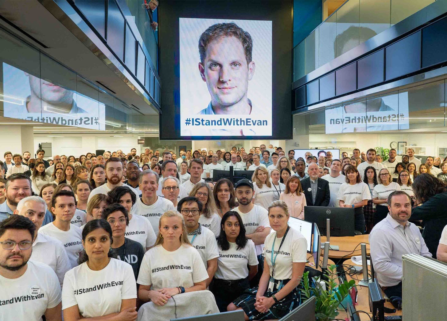 A crowd in the Wall Street Journal newsroom gathers around a screen projecting a headshot of reporter Evan Gershokovich and the hashtag #IStandWithEvan