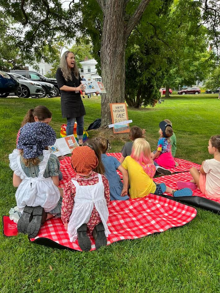 Photo of me (Julie Falatko) Reading Help Wanted One Rooster) to some children who are sitting on red checkered ground cloths, outside, under a tree. It's very picturesque. This area was called the "Storytime Nook," which gives you a sense of how picturesque and cute it all was.