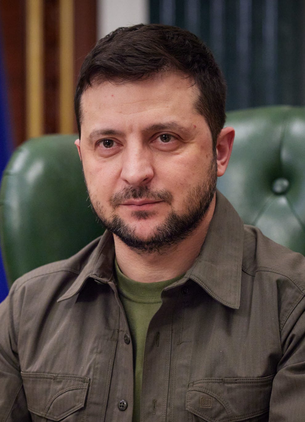 Zelenskyy in 2022, wearing green fatigue like outfit and a beard; he is seated and the picture is from chest up.