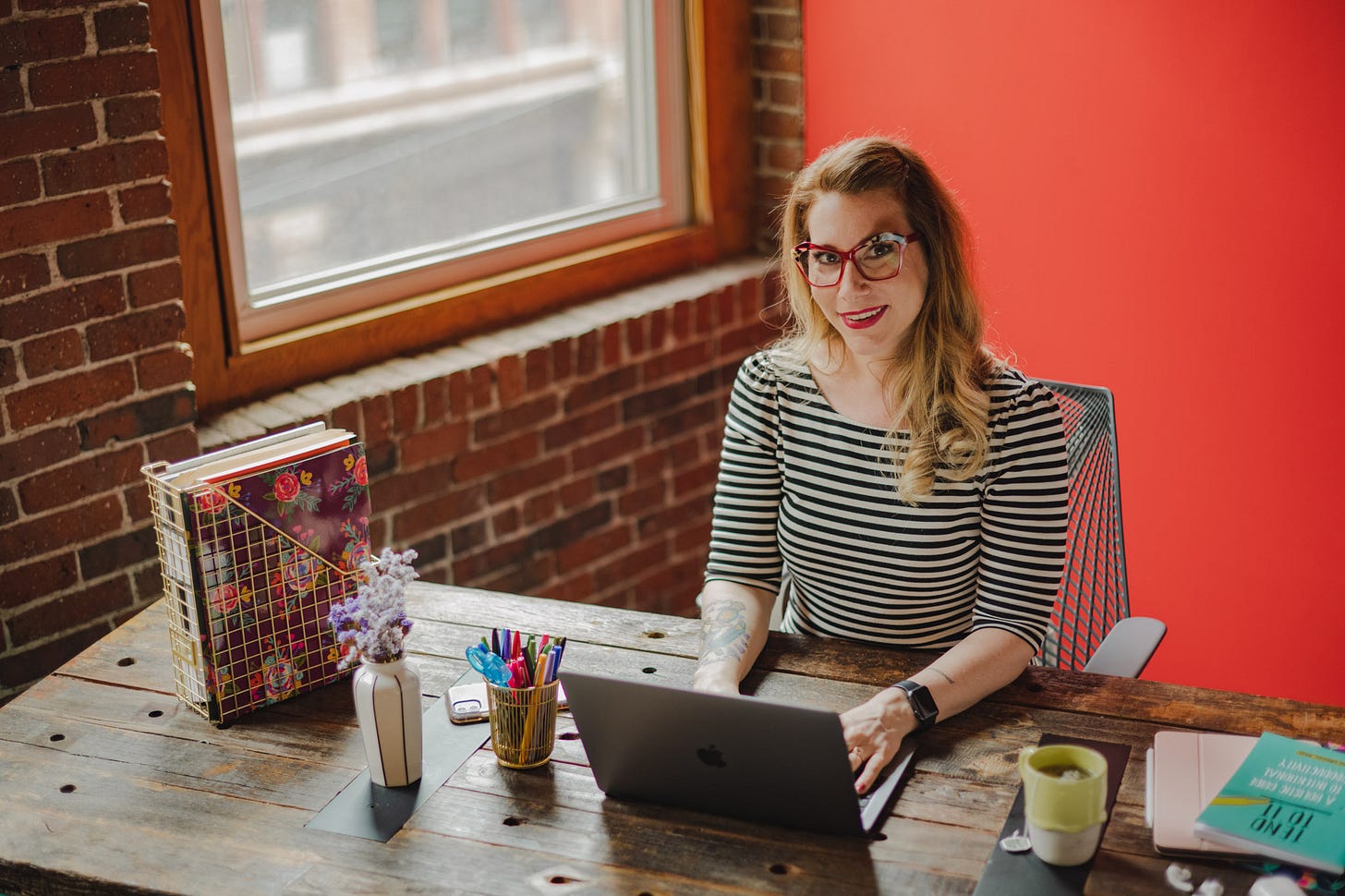 Dr Kate is a white woman sitting at a wooden desk with a laptop, pens, books and a cup of tea on the desk